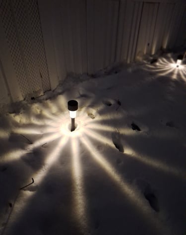 Solar pathway lights casting a starburst pattern onto white snow along a garden fence at night.