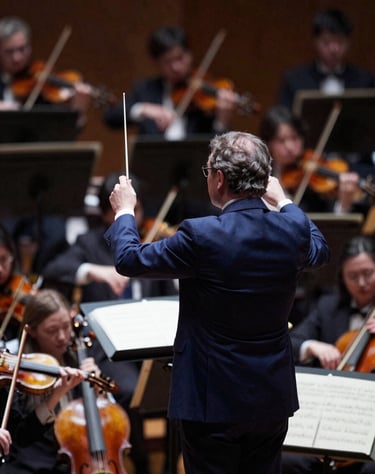 A dramatic shot of the orchestra pit from above, with the conductor's baton raised. Colors include deep #2B2D42 and orchestral highlights in #C8B8B8.