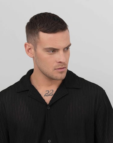 Portrait d’un modèle masculin en chemise noire sur fond gris clair, regard tourné sur le côté, ambiance studio épurée.