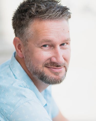 Portrait d’un comédien souriant en chemise claire, posé en extérieur avec un fond flou, idéal pour un book professionnel.