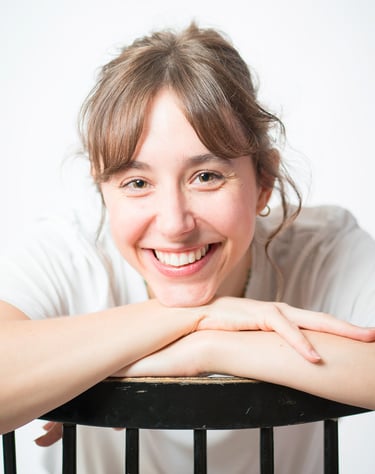 Portrait d’une comédienne souriante en studio, en t-shirt blanc, appuyée sur un dossier de chaise noire, sur fond blanc lumin