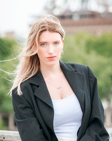 Portrait d’une modèle en veste noire, posant en extérieur avec un regard intense, sur fond flou de verdure et d’architecture 