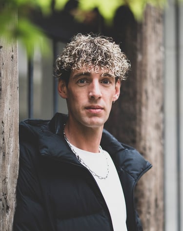 Portrait d’un modèle au regard intense, posé près d’un mur en bois, avec une lumière naturelle et un fond de feuillage flou.