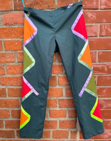 Dark teal pants with colorful geometric patchwork hanging against a rustic red brick wall.