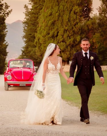 Sposi che camminano per mano lungo un viale alberato, sorridenti davanti alla loro auto d’epoca
