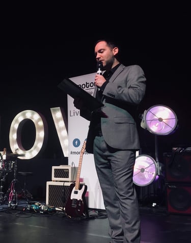 Alex hosting an awards function in a grey suit holding a clipboard
