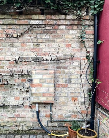 An electricity box attached to a garden brick wall painted in camouflage brick pattern.
