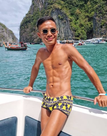 A male outcall masseur in pattaya standing on a boat wearing swim wear with the sea and an island in the background