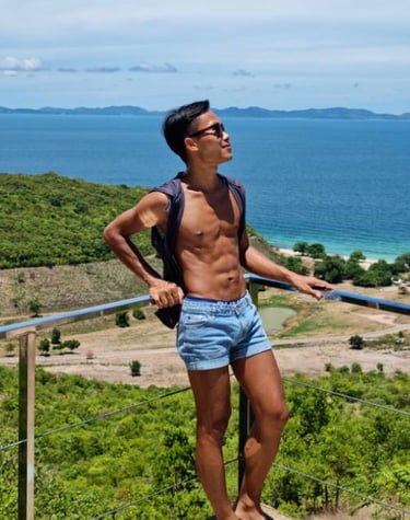 Man wearing blue shorts and no top standing on a viewpoint with sea and an island in the background.