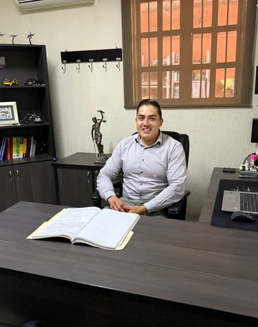 A smiling professional lawyer sitting at a dark wood desk in an organized office with law books and a laptop.