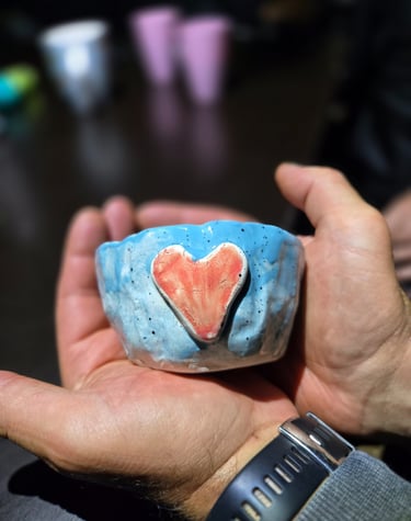 Handmade light blue ceramic bowl with a red clay heart being held in a pair of open hands.