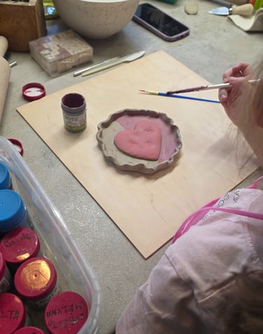 A young child painting a pink ceramic heart dish with a brush during a pottery craft workshop.
