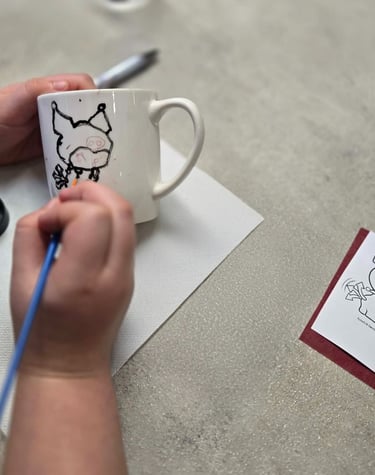 A person hand-painting a cartoon character onto a white ceramic coffee mug with a blue paintbrush.