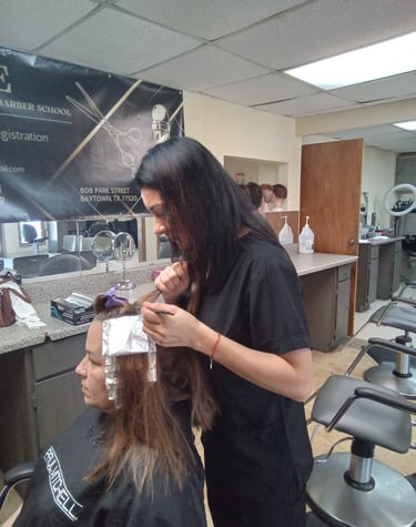 A student at a beauty school applying hair highlights with foil to a client in a salon setting.