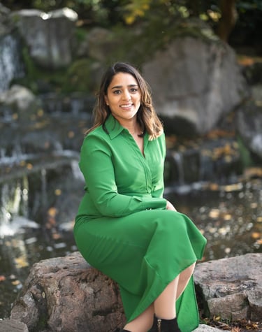 A photo of Anita wearing a green dress. She is sat on a rock with a water feature in the background.