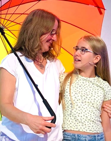 eine Frau mit einem Kind, die beide Headsets tragen und singen unter einem bunten Regenschirm