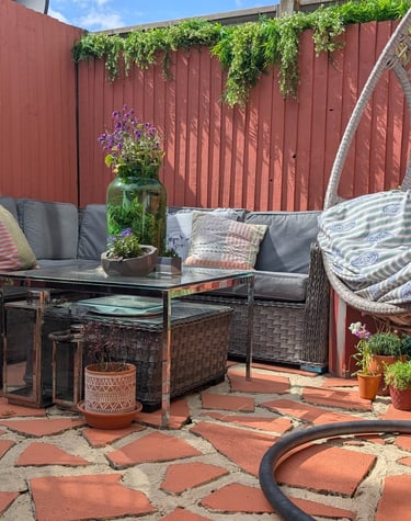 A cozy patio with a grey wicker corner sofa, hanging egg chair, and garden plants on terracotta tiles.