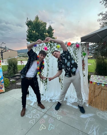 Two men in Hawaiian shirts make a heart shape at a tropical theme outdoor party.