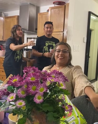 Friends celebrate at a house party with purple flowers, snacks, and drinks in a kitchen.