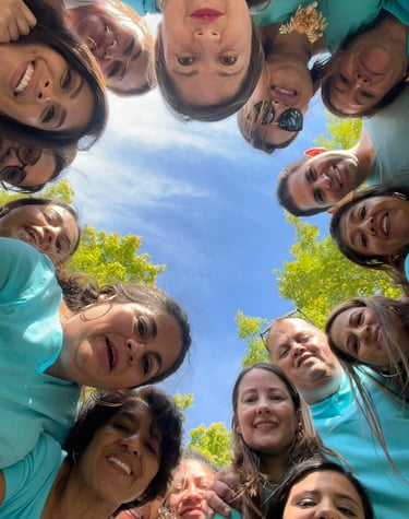 A diverse group of smiling people in teal shirts posing for a low angle circle selfie outdoors.