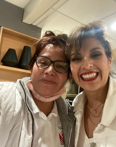 Two smiling women taking a selfie indoors wearing white jackets and casual accessories.