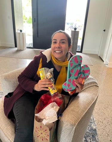 Smiling woman sitting in a chair holding a gift bag, watermelon print flip-flops, and small toys.