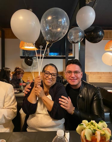 Smiling woman holding white balloons sitting with a man in a leather jacket at a birthday dinner party.