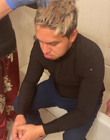 Professional applying blonde hair bleach to a man's short hair in a home bathroom setting.