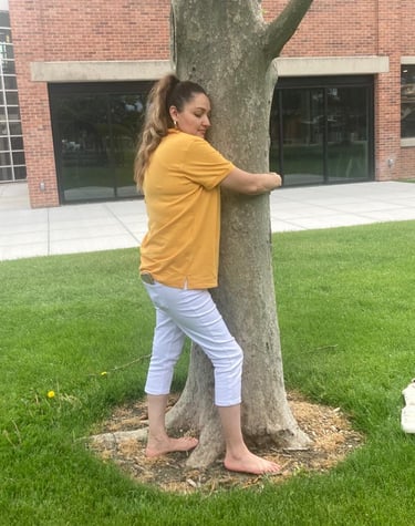 A barefoot woman in a yellow shirt and white pants hugging a large tree on a green lawn.