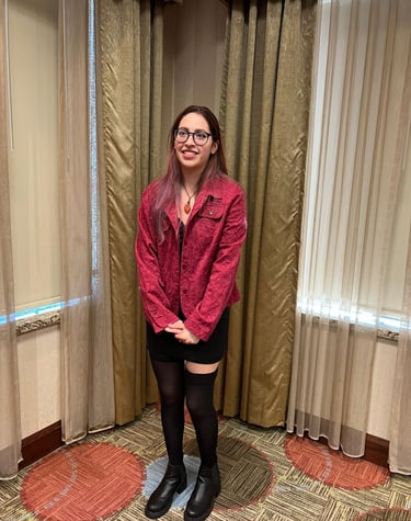 A smiling woman with glasses wearing a red textured jacket, black skirt, and boots standing in a room.