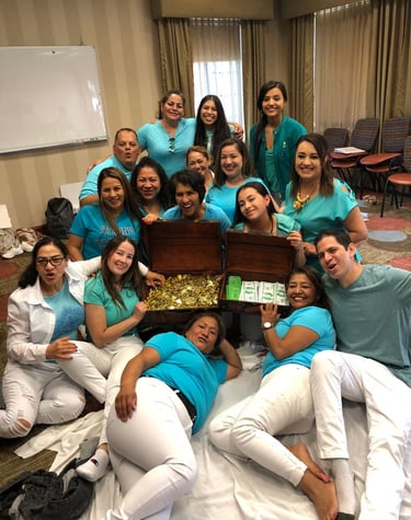 A cheerful group of 18 women and 2 men, dressed in blue and white, pose together around a treasure chest overflowing with