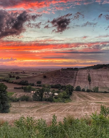 A vibrant sunset over rolling hills and rural farmland in the Italian countryside.