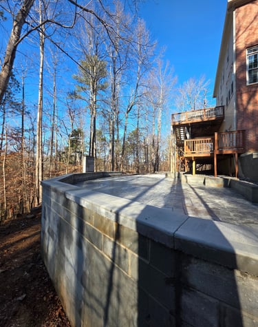 Custom stone patio and retaining wall under construction behind a brick home with a wooden deck.
