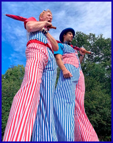 Two circus performers on stilts wearing red and blue striped trousers and holding umbrellas.