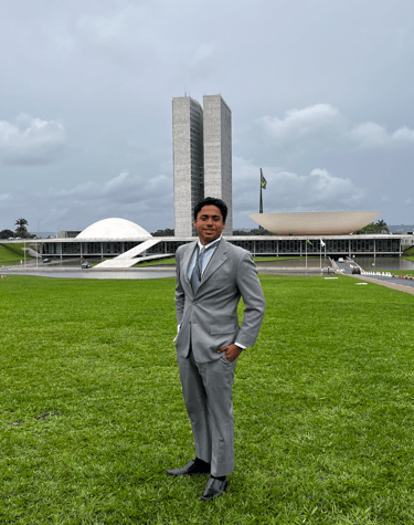 Jovem estudante vestido com terno cinza posando em frente ao Congresso Nacional em Brasília, represe