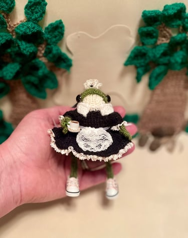 a person holding a small knitted frog dressed as a french maid in a hand