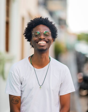 Homem preto e magro, de cabelo crespo. Usa óculos de armação fina e redonda. Tem o sorriso largo.