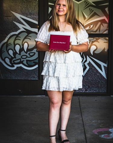 A smiling blonde graduate in a white tiered dress holds a red diploma, celebrating her achievement!