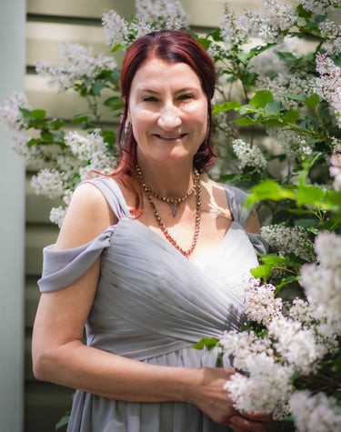 Smiling woman with auburn hair in a grey dress posing by white lilac bushes.