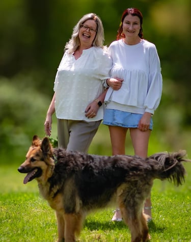 Two smiling women standing together in a sunny park with their large long-haired German Shepherd dog.