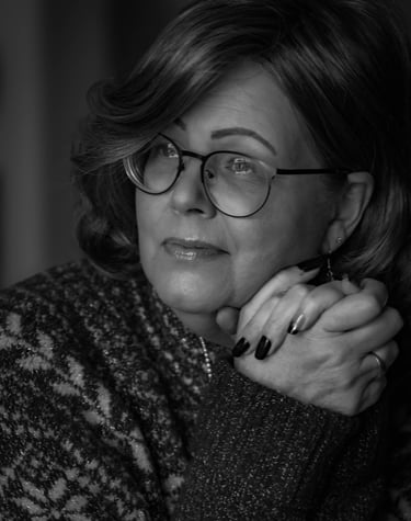 Black and white portrait of a thoughtful, mature woman with eyeglasses, resting her chin on clasped hands.