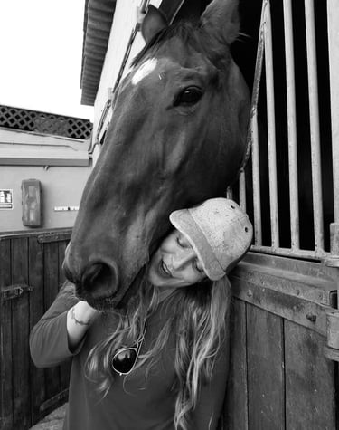 A smiling woman wearing a baseball cap leans against a brown horse standing in a stable stall.