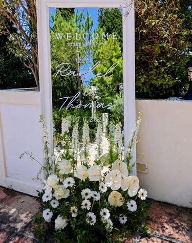 A white-framed mirror wedding welcome sign for Rachel and Thomas decorated with white flowers.