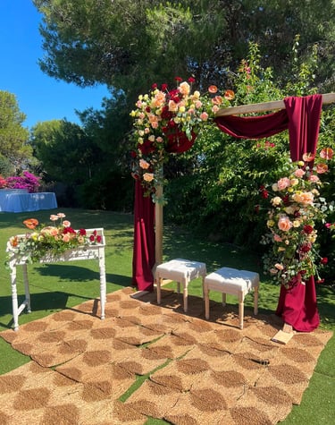 Outdoor wedding ceremony setup with a floral wooden arch, red drapery, two white stools, and a woven rug on the grass.