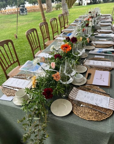 Long outdoor banquet table set with green linen, woven placemats, and a colorful floral centerpiece.