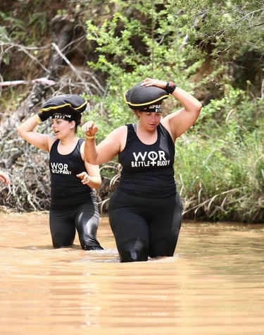 Two women participating in a Spartan Race obstacle course carry sandbags while wading through muddy water.