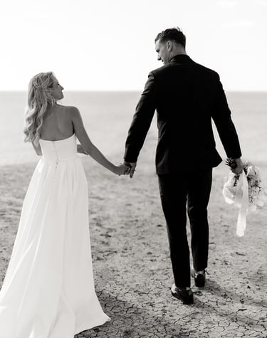 a bride and groom holding hands and walking down a beach