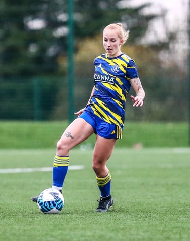 "Female soccer player in blue and yellow uniform dribbling on a field during a match.