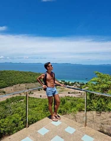 Male therapist wearing blue shorts and no top standing on a viewpoint with sea and an island in the background.