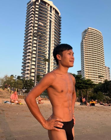 Man standing confidently on Pattaya Beach at sunset, with tall modern buildings and palm trees in the background.